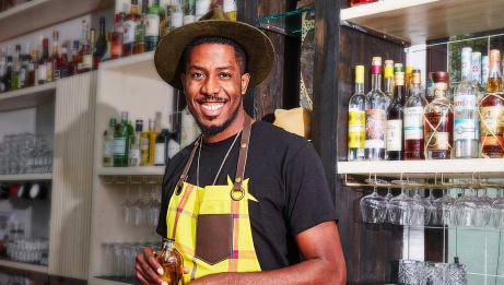 Paul Toussaint se tient devant un bar, un verre à la main. Il regarde vers la caméra et sourit. Paul porte un tablier jaune et un chapeau à larges bords. © Darwin Doleyres