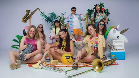 Vibrant group of musicians playing brass instruments, donned in summer clothing amidst a beach scene with toys, an outdoor cooler, beach towel, and faux plants.