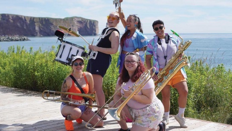 Five people in summer attire holding musical instruments, joyfully posing on a boardwalk with an ocean and rocky cliffs in the background, radiating smiles and capturing a moment of fun.