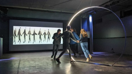 Four adolescents standing hand in hand around a giant ring light, with a distorted projection of their image cast onto the screen in front of them.