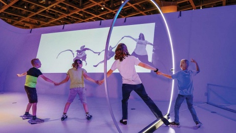 Four children standing hand in hand within the glow of a giant ring light, with a distorted projection of their image cast onto the wall in front of them.