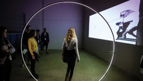 Girl standing in the center of a giant ring light, with her distorted image projected onto the screen to her right. On her left, five other people are not visible in the projection.