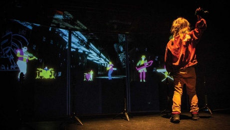 Young girl with pigtails seen from the back, hand raised and joyfully dancing while looking at three screens displaying neon images of people and instruments