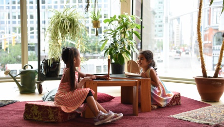 Two little girls sitting on floor pillows, facing each other with a table between them. One girl holds a book, while the other holds a candle. They appear happy, surrounded by plants.