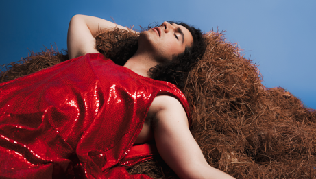 Jeremy Dutcher wearing a sparkly red garb lounging on a hay pile