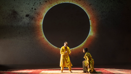 One person standing and one kneeling, both wearing yellow robes with a projected image of a solar eclipse behind them. 