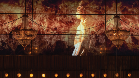 Two chandeliers hanging with a projection in the background of a topless man with fire surrounding him.