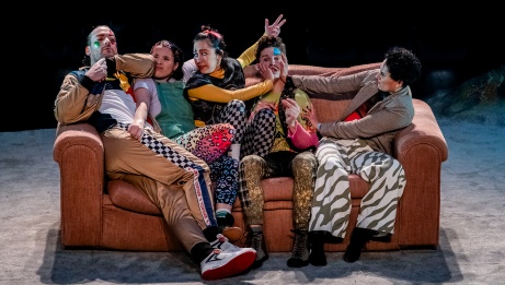 Five performers struggle to get a seat on a sofa too small to accommodate them all comfortably
