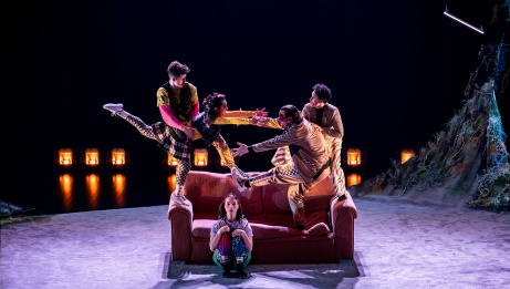 Four performers are standing on a sofa, mid-jump. A fifth performer is crouched on the ground in front of the sofa.