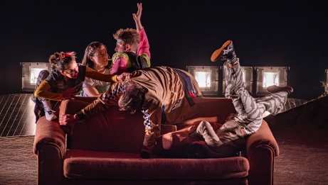 Five performers struggle to get a seat on a sofa too small to accommodate them all comfortably.