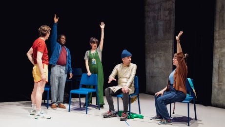 Five characters gather on stage, surrounded by a theatrical setting with brightly colored chairs and clothing. Three of them raise their hands to speak, while two others observe them closely.