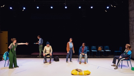 Six characters are scattered on stage, immersed in a theatrical atmosphere. One withdraws, engrossed in reading a text, another passionately expresses themselves, capturing the attention of some, while others turn their gaze across the room.