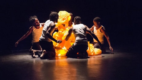 Five dancers in white camisoles and black pants kneel in a circle around a strange sculpture of orange plastic bags. 