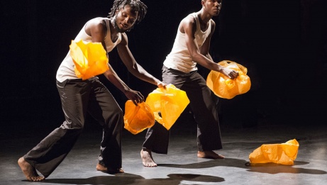 Two dancers in white camisoles and black pants juggle orange plastic bags. 
