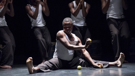 A dancer sits on the floor, legs spread. He plays with a ball. Behind him, standing, dance 4 other dancers dressed in white camisoles and black pants. 