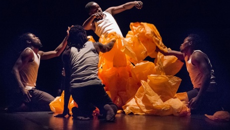Four dancers in white camisoles and black pants kneel in a circle around a strange sculpture of orange plastic bags.
