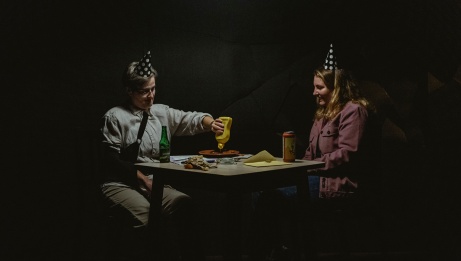In a softly lit room, two seated individuals wear festive hats. One of them pours mustard onto a plate.