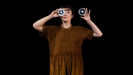  A young woman is standing and holding reels of film in front of her eyes. She wears a brown jumpsuit and pink slippers.