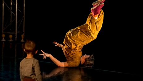 The dancer is lying on her back, legs to the sky. She wears a brown jumpsuit and pink slippers. A child, from behind, looks at her.