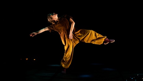 A dancer is standing, moving, with one leg raised. She wears a brown jumpsuit and pink slippers.