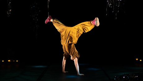 The dancer balances on her hands with her legs apart. She wears a brown jumpsuit and pink slippers.