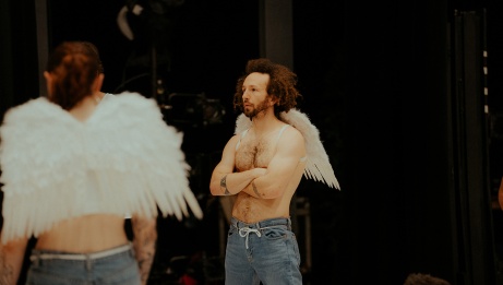 A shirtless man adorned with white wings attached to his body. By his side, a slightly more blurred figure is also equipped with wings