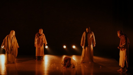 In a room bathed in a yellow glow, four individuals draped in fabrics with their faces completely concealed observe a figure on the floor