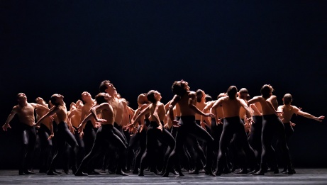 A large group of dancers on stage in black tights and skin toned tops in a coordinated arched gesture towards the light.