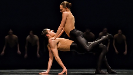 Two dancers in a pose on stage, one straddling the other, with five performers in the background in dim light. 