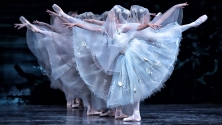 Ballet dancers in white costumes with veils perform on stage, posing with one leg and both arms extended, facing in opposite directions. 