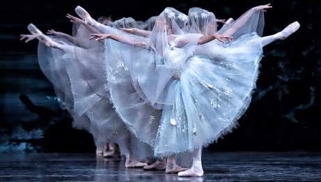 Des danseuses de ballet en costumes blancs et voilées se produisent sur scène, posant avec une jambe et les deux bras tendus, dans des directions opposées. 