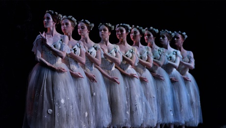 A straight line of ballet dancers in white costumes and floral headpieces, positioned on stage with arms crossed against their bodies. 