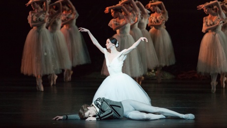A ballerina in a white tutu kneeling behind a male dancer lying at her feet, with three lines of dancers in the background.