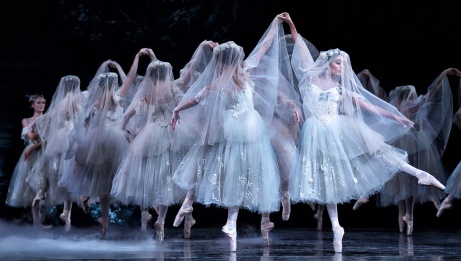 Ballet dancers in white costumes with veils on pointe clasping hands above their heads. 