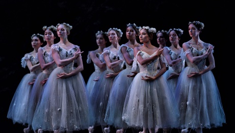 A line of ballet dancers in white costumes and floral head pieces pose with folded arms and tilted necks. 