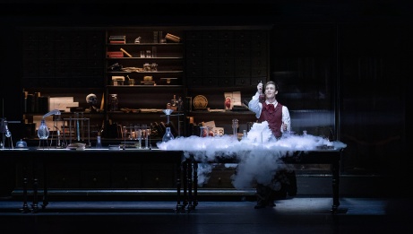 A male dancer stands in his laboratory holding a smoky potion. 