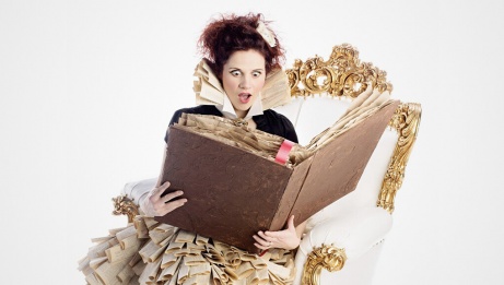 Luana is sitting in a white armchair with gold details, reading a storybook. She's wearing a theatrical costume and her facial expression is one of surprise at what she's reading.