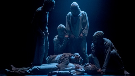A group of performers in hoodies stand around a figure lying on the stage, under dim blue lighting, evoking a somber mood.