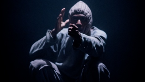 A performer dressed in a grey hoodie crouches on stage with one finger pointing forward. 