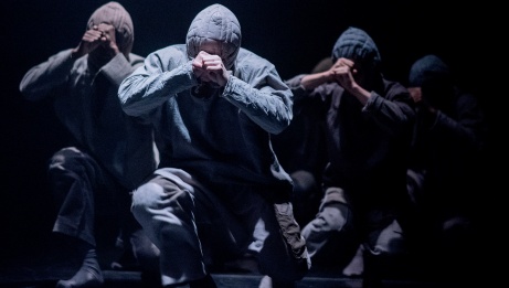 4 dancers in grey hoodies kneel on a dark stage with closed fists positioned at their foreheads. 