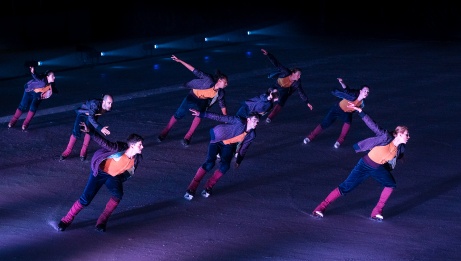 9 performers on skates in purple costumes move in formation across the ice. 