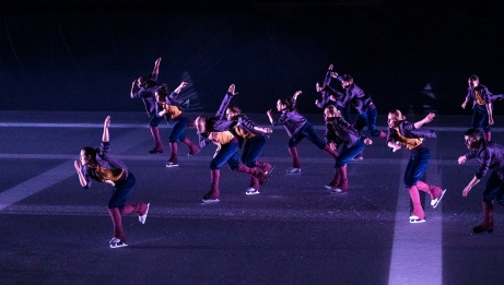 12 performers on skates in purple costume move in a group formation across the ice.  