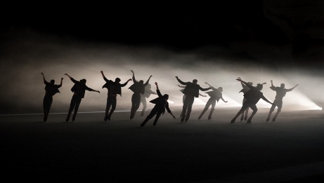12 performers on skates back lit with white light and fog, arms extended at their sides. 
