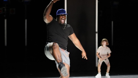 A man wearing shorts and a snapback hat is in a stomping position with his mouth wide open. In the background is a young dancer in a pose. 
