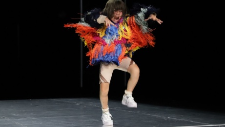A young dancer in a colourful jacket with frills is in a stomping position with both arms out at her side. 