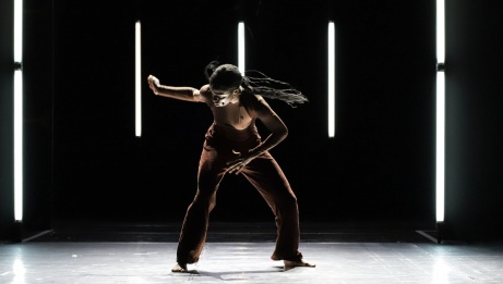 A dancer with braids leaning over mid movement on stage in front of 5 lines of bright lights. 