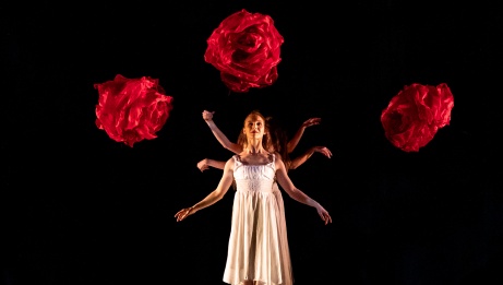 A performer in a white dress appears to have six arms, standing in front of three large floating red roses. 