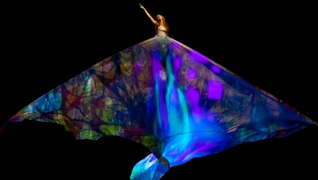 A dancer draped in a large, colorful wing-like costume extends one arm upwards on a dark stage. 