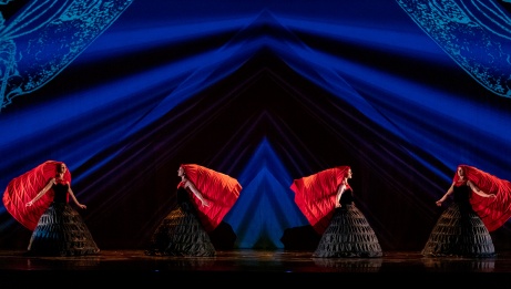 4 dancers in black dresses with wide skirts, and large red headpieces, stand against a blue geometric backdrop. 