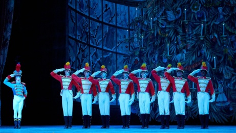 A row of dancers lined up in matching red soldier uniforms with one hand at their foreheads. Off to the left, is a toy soldier of similar height wearing a blue soldier uniform. 
